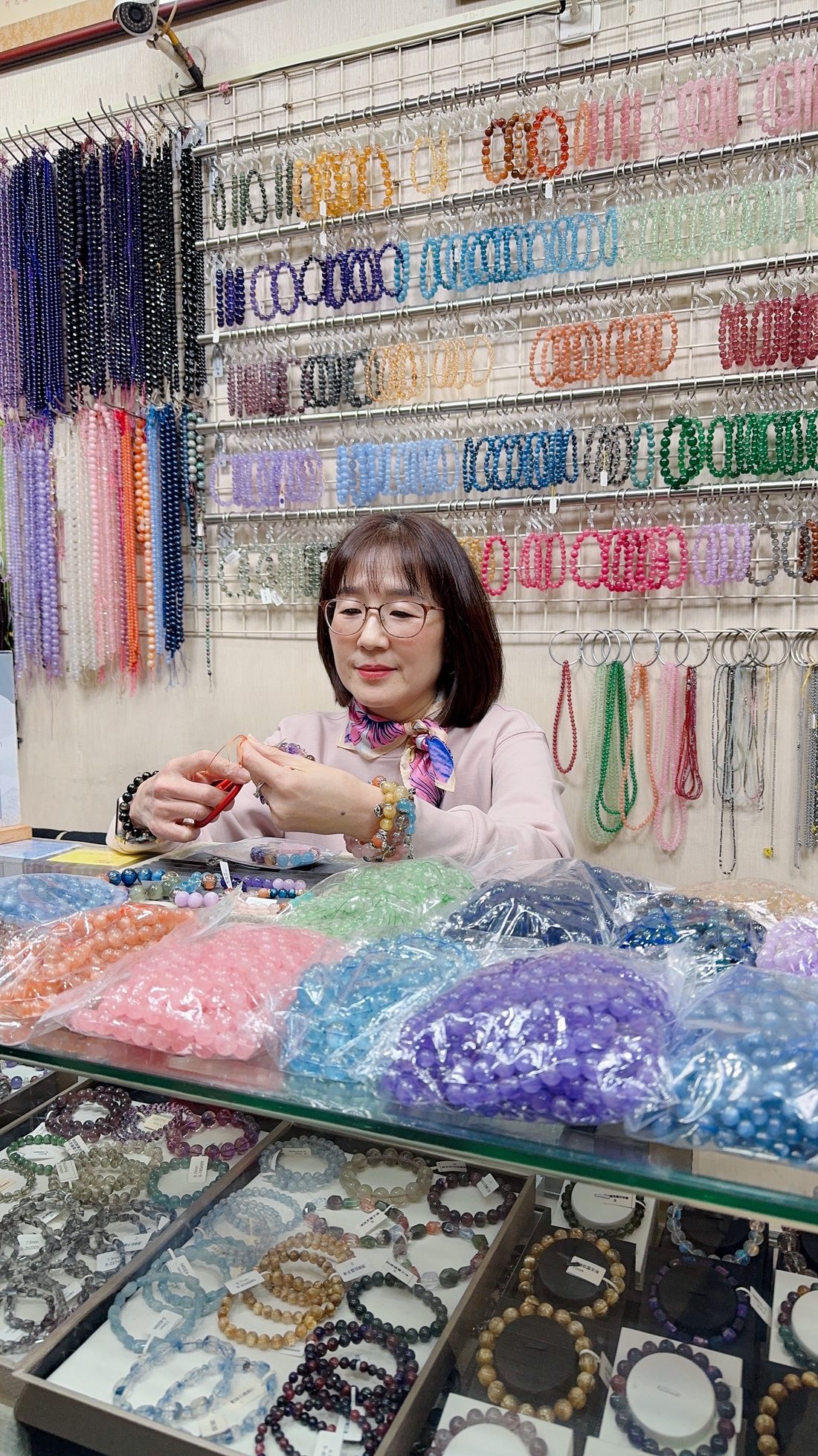 紋君老師坐在店內工作檯前串製手鍊,身後整面牆掛滿各色水晶珠串,桌上是分裝好的散珠,玻璃櫃裡是貼好標籤的成品手鍊