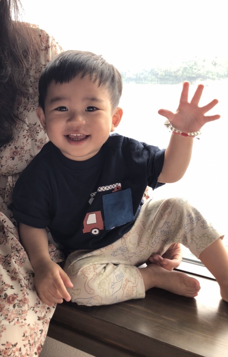 男孩坐在海邊的木桌上揮著手，抬起的手腕上戴著寶寶生命密碼手鍊