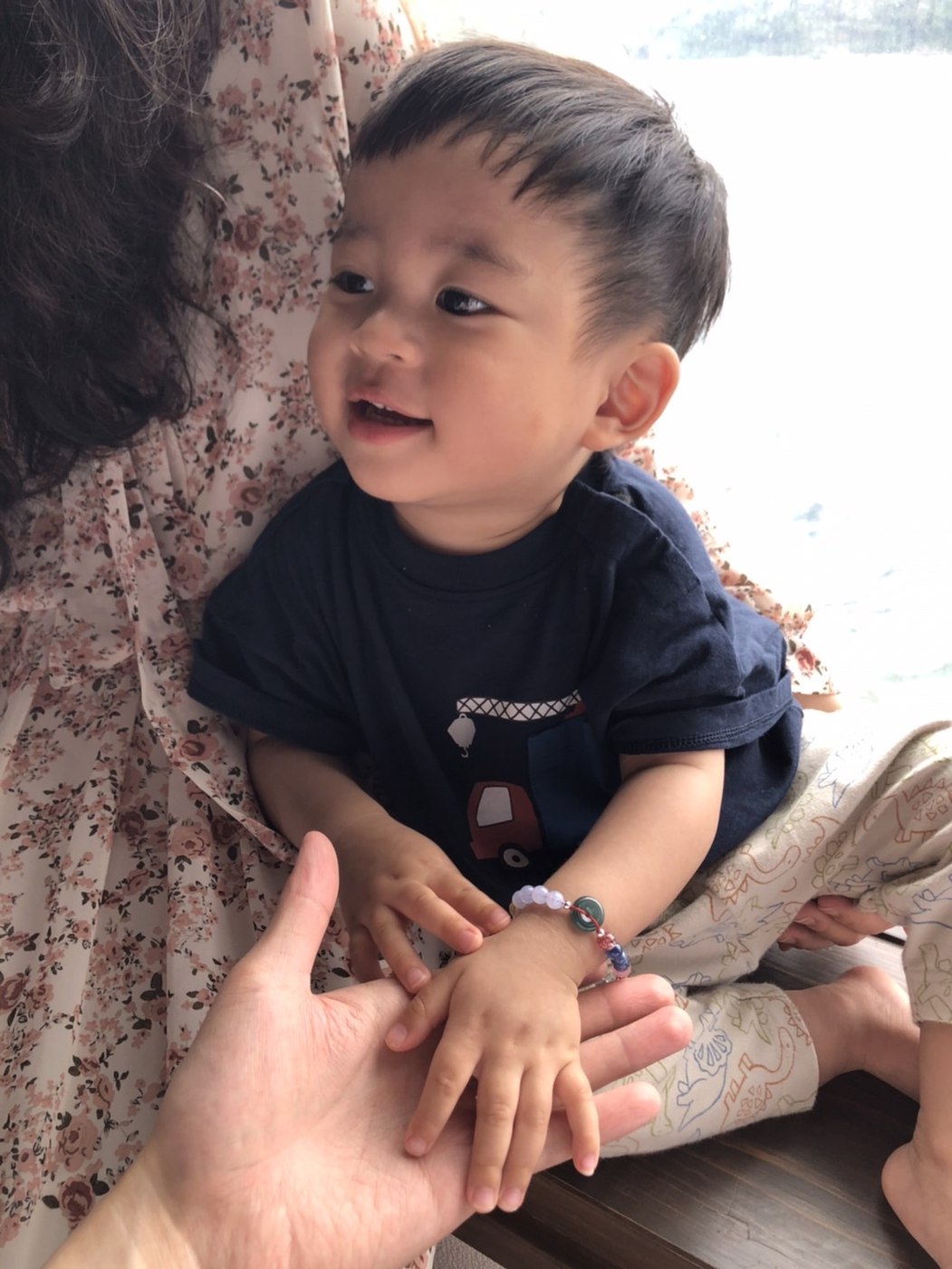 一歲多的男孩坐在大人身邊，手腕上戴著寶寶生命密碼手鍊，中間是一顆藍水翡翠平安扣